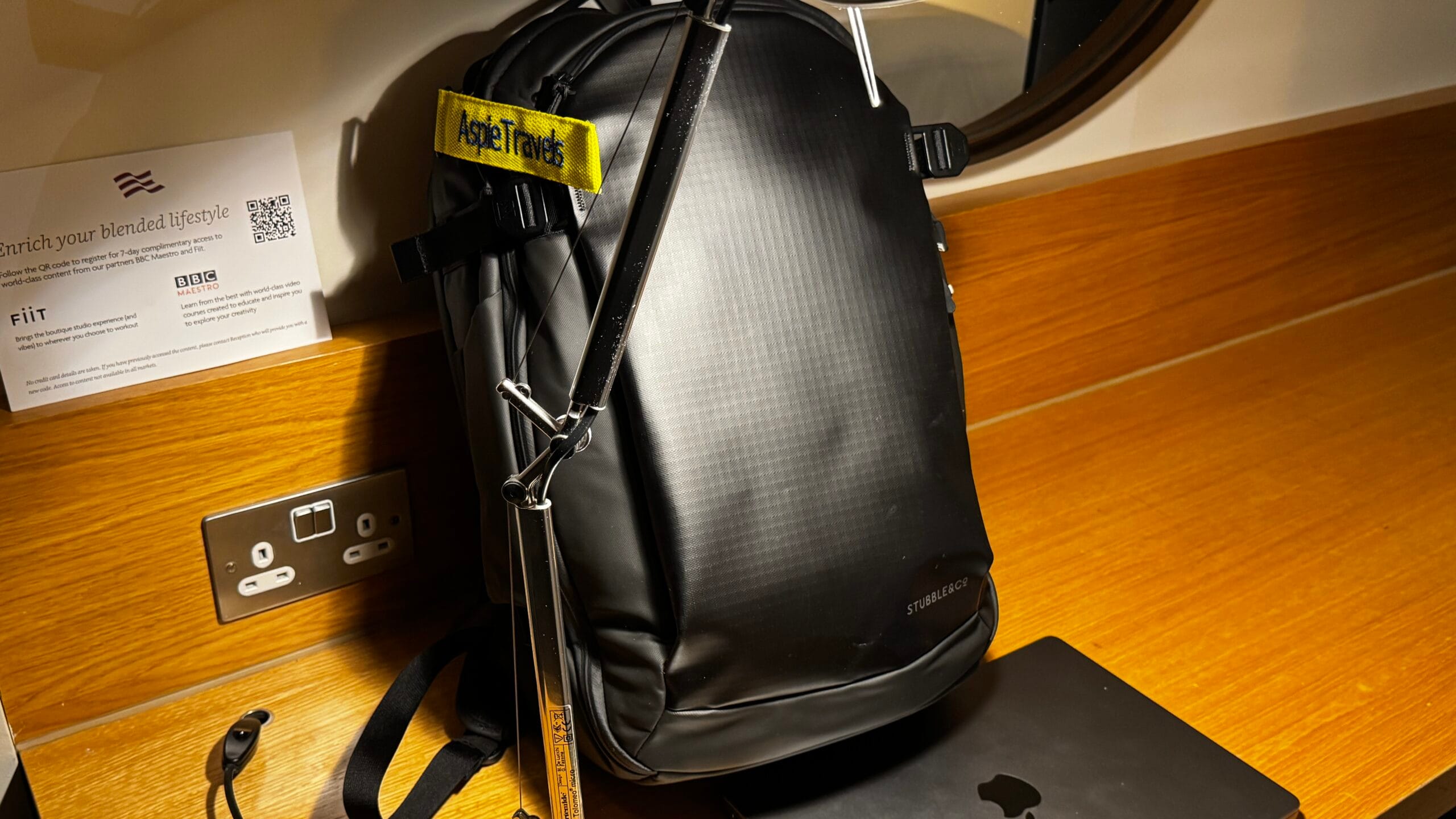 Stubble 30L Hybrid backpack set up on a hotel desk with charging cable and AspieTravels luggage tag attached