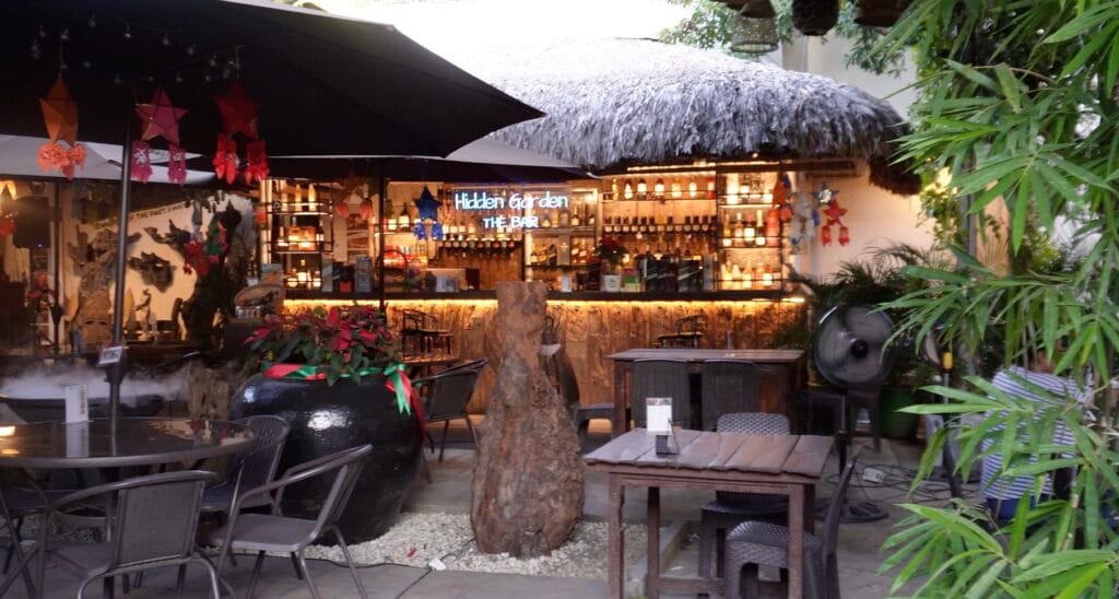 Hidden Garden The Bar outdoor tropical setting with thatched roof, warm lighting and a fully stocked drinks bar surrounded by plants