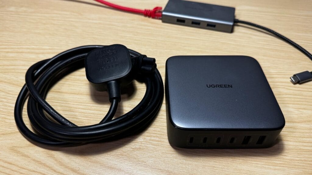 Close-up of a UGREEN multiport charger showing four USB-C ports and two USB-A ports beside a fused UK plug on a wooden surface.