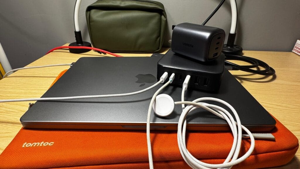 Closed MacBook on an orange Tomtoc sleeve with multiple white charging cables connected to a stacked black UGREEN power hub and charger on a wooden desk.