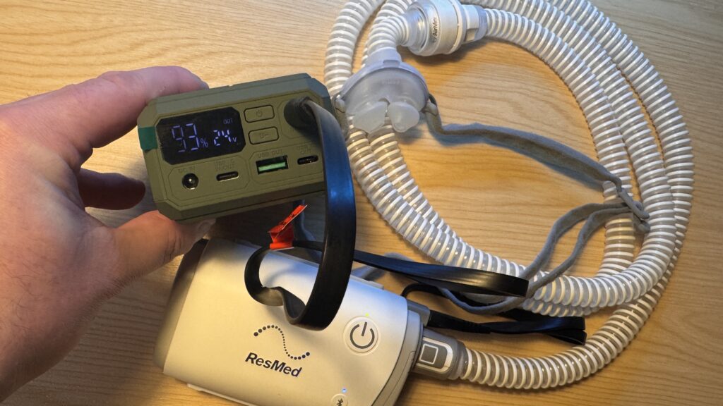 Hand holding a travel battery showing 93% charge and 24V output while powering a ResMed AirMini